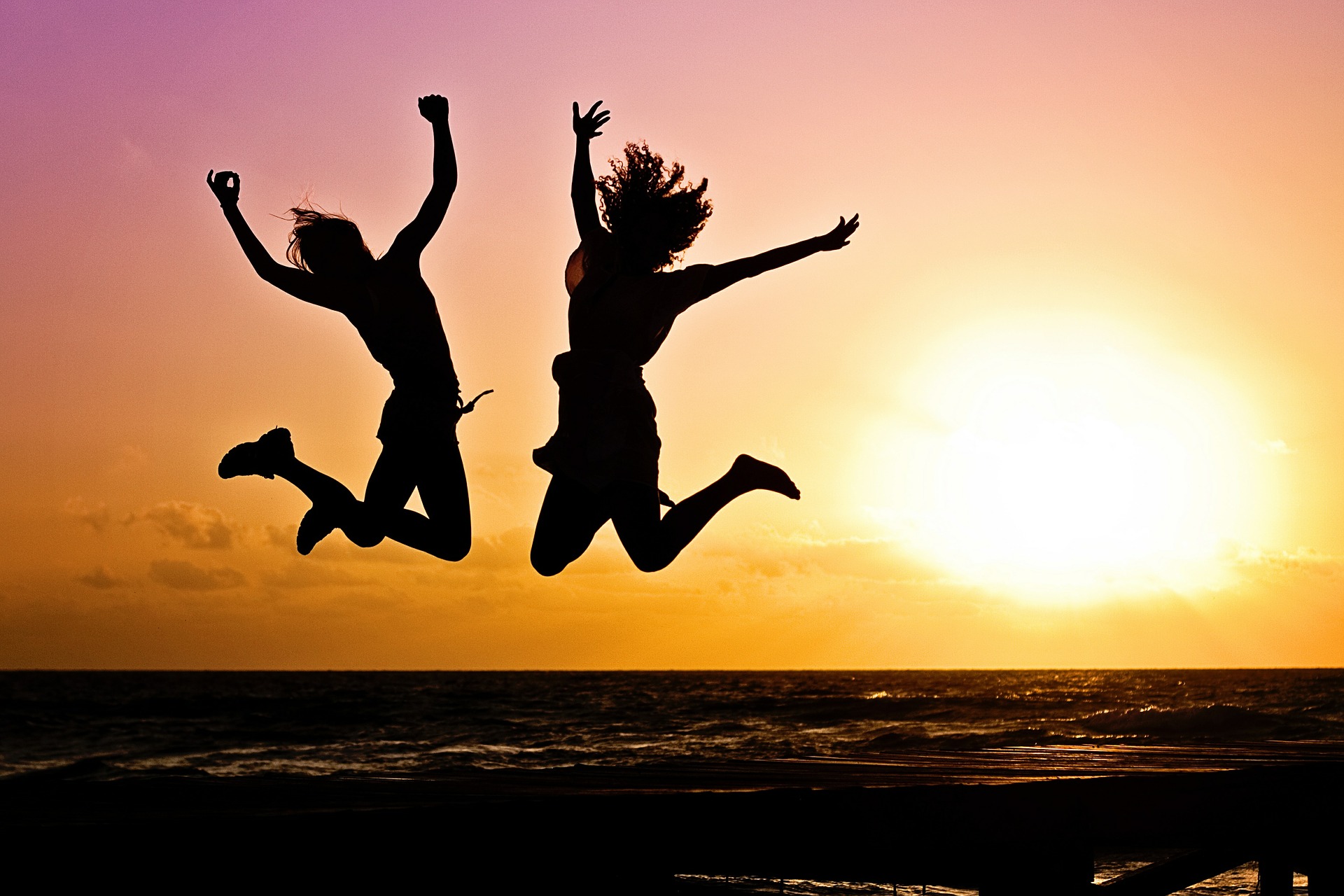 Two silhouettes of people jumping high with their arms aloft in front of a sunset backdrop.