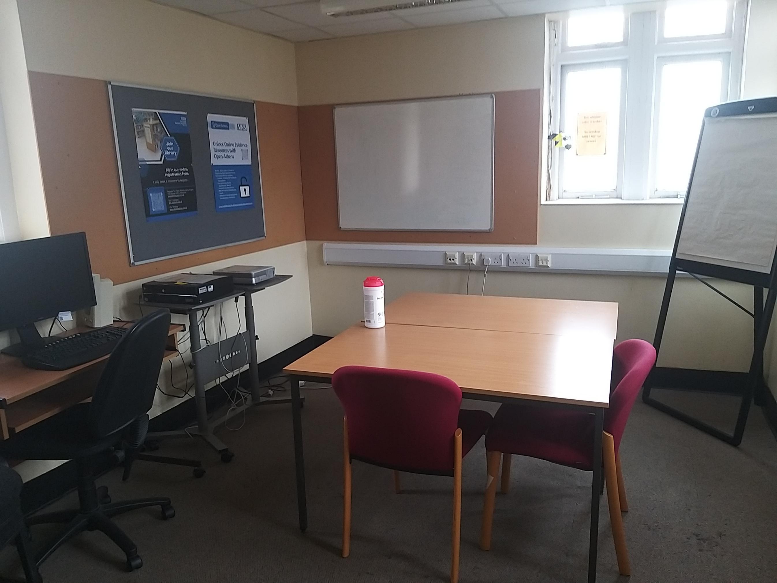Photo of square desk with chair around two sides,wall mounted whiteboard and flip chart and projector connected to PC.